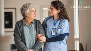 A female senior assisted living nurse wearing blue scrubs smiling while steadying her senior female patient by holding her arm