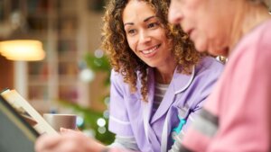 A senior woman sitting with her female nursing assistant looking down at an iPad