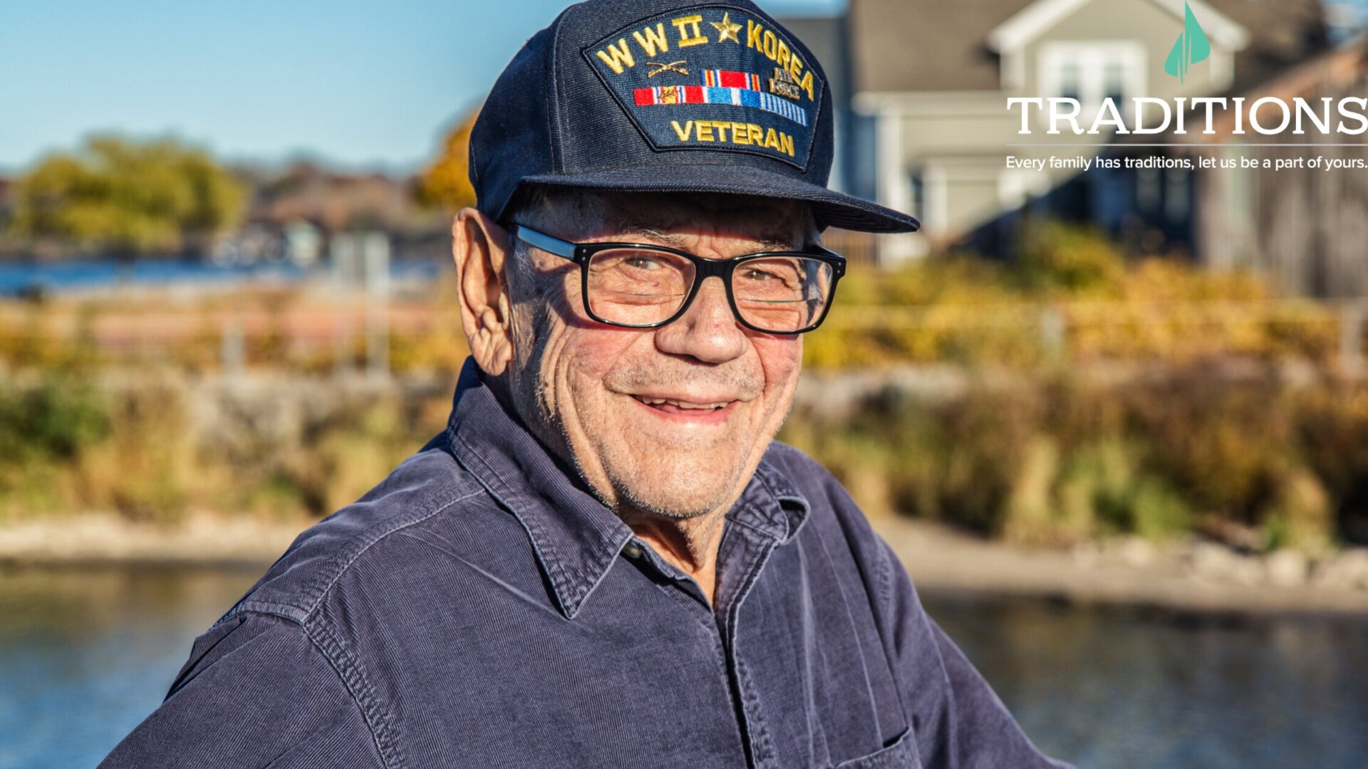 Photo of a male veteran wearing his veteran hat and black glasses as he stands in front of a river. The Traditions Management logo is in the top right corner of the image.